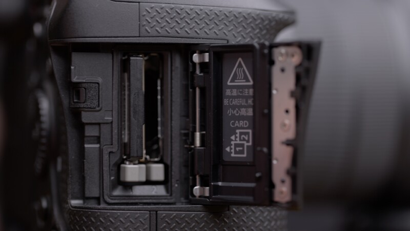 Close-up of an open camera slot showing two memory card slots. The card compartment cover is open, revealing a caution symbol and instructions. The camera body has a textured grip surface.