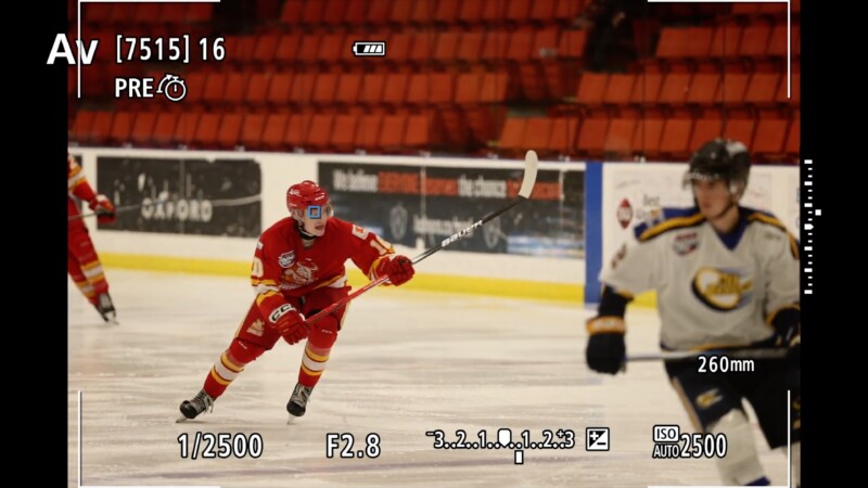 A hockey player in a red uniform skates across the rink in mid-action, holding a stick. Another player in a white uniform is nearby. The scene is framed by a camera’s viewfinder display, showing settings like ISO and shutter speed.