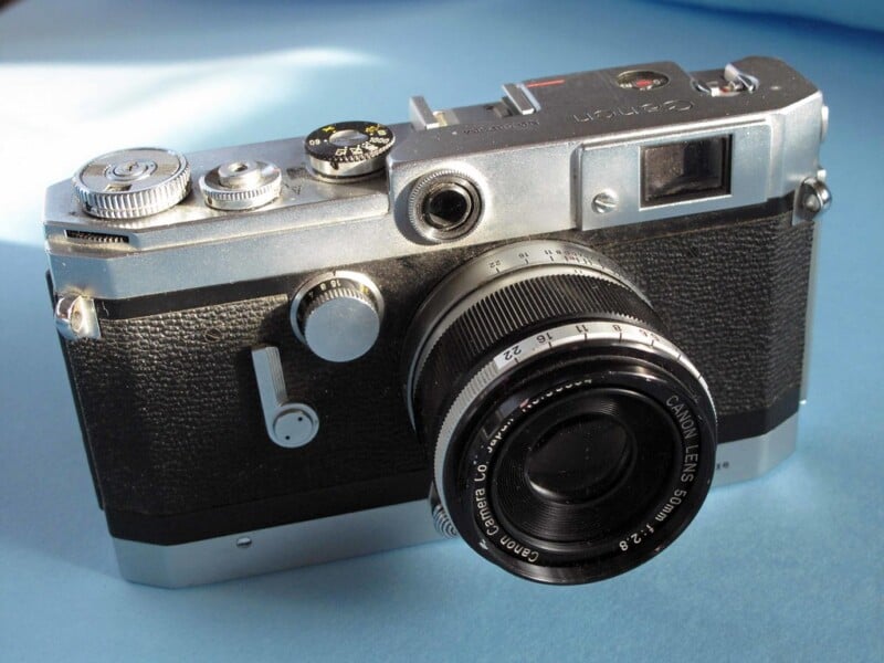 A vintage Canon rangefinder camera with a black and silver body sits on a light blue surface. The camera features a prominent lens and several dials and buttons on the top.