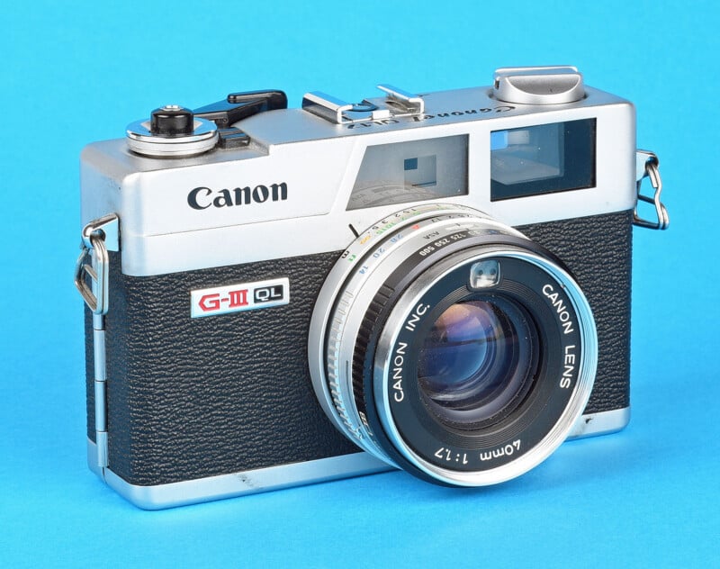 A vintage Canon G-III QL camera is displayed against a blue background. The camera features a black textured body with silver accents and a prominent Canon lens.