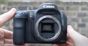 A Canon EOS 10D digital camera is held by two hands against a blurred brick wall background. The lens is removed, revealing the camera’s sensor and internal components. The camera's brand name and model are clearly visible on the front of the body.