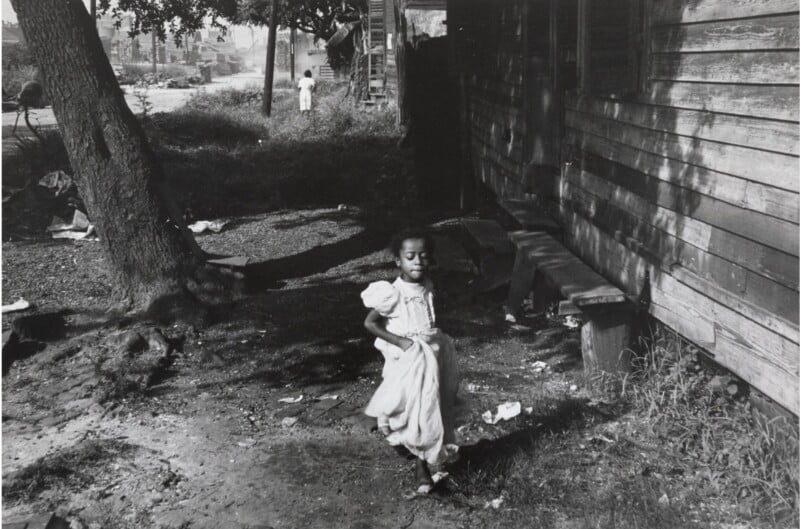 A young girl in a dress runs through a dirt yard beside a wooden house. A large tree stands nearby, and a person is visible in the background. The environment looks rustic and weathered, with scattered debris and greenery.