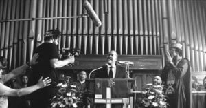 A black-and-white photo shows a man speaking at a church podium, surrounded by people. A large microphone hangs from above, and two men stand to his left—one holding a camera and the other in clergy attire. Floral arrangements decorate the scene.