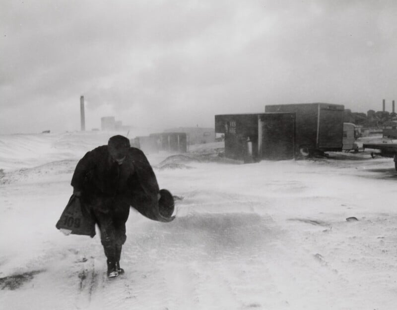 A person walks through a snowy, industrial area, bundled in heavy clothing against the wind. They carry a bag and shield themselves with an arm. Industrial buildings and trucks are partially obscured by snowdrift and fog in the background.