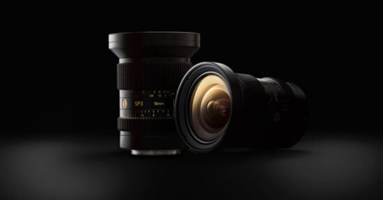 Two black camera lenses displayed on a dark surface with a black background. One lens is standing upright, while the other is positioned horizontally, showing the golden interior of the lens. Both lenses feature various markings and numbers.