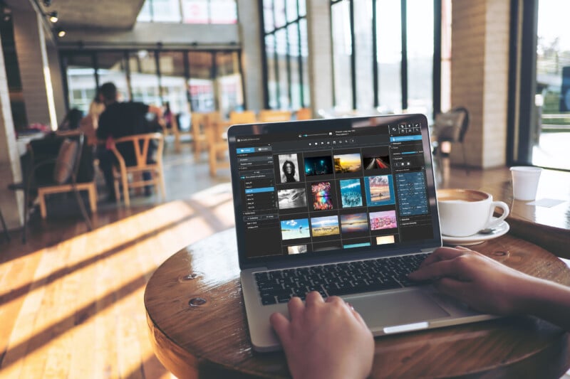 A person using a laptop at a wooden table in a cafe, with a coffee cup nearby. The screen displays a photo editing software interface. The cafe has large windows and several empty chairs and tables in the background.