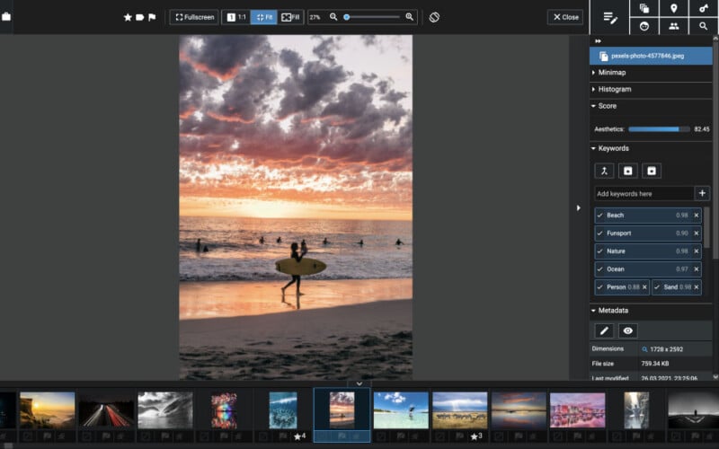 A surfer carrying a surfboard walks along the beach at sunset. The sky is filled with dramatic clouds reflecting vibrant orange and pink hues over the tranquil ocean, with several surfers in the water.