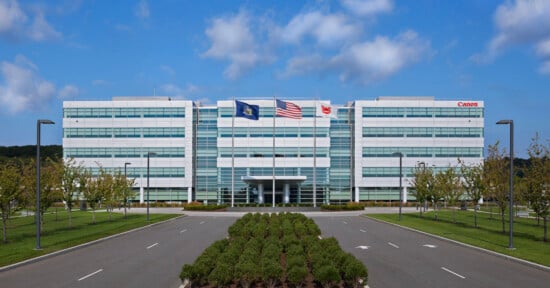 A modern office building with multiple floors, featuring a glass entrance and flags of the state, the United States, and a corporate logo above the main entrance. There are well-maintained green bushes and a clear driveway leading to the entrance, set against a blue sky with scattered clouds.