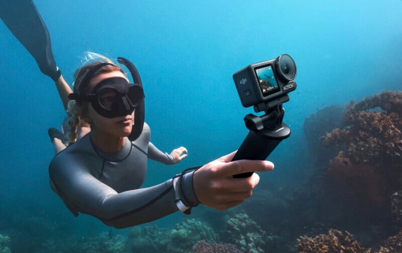 A person wearing a wetsuit and snorkeling gear swims underwater, holding a camera on a handle. Coral and marine life are visible in the background, with blue ocean water surrounding them.