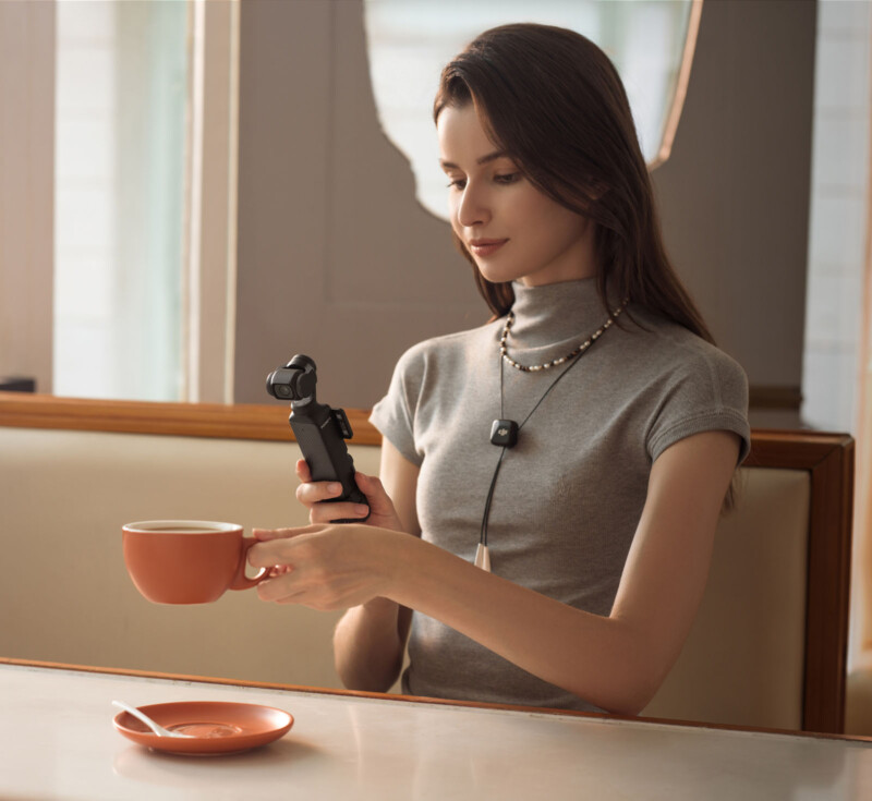 A woman sitting at a table holds a camera device while looking at it. She is wearing a gray turtleneck and a necklace. A cup of coffee sits on a saucer in front of her on the table. The setting appears to be a cafe.