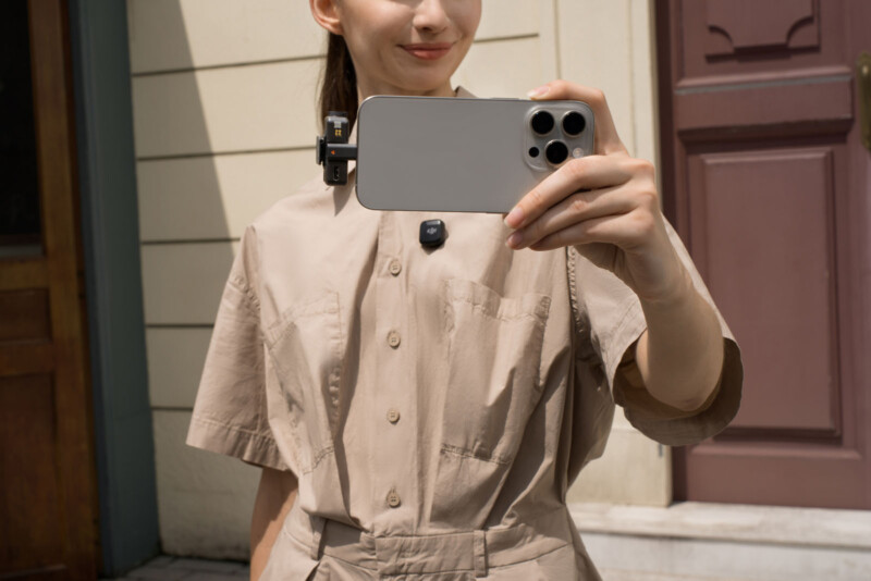 A person wearing a beige outfit is taking a selfie with a smartphone. The phone is held in their right hand, with a clip-on mount visible. They are standing in front of a building with a cream-colored wall and a wooden door.