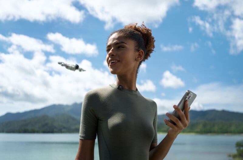 A person stands by a lake holding a phone, with mountains and clouds in the background. A small drone flies nearby. They are looking up and smiling, wearing a green shirt. The setting is sunny and peaceful.