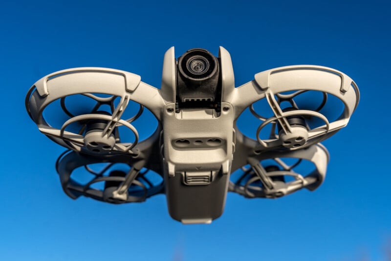 A small drone with four propellers is flying against a clear blue sky. The drone's camera is visible on the front, and the propellers are encased in protective guards.