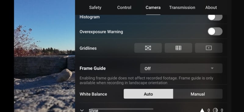 Camera settings interface on a digital device screen displaying options for histogram, overexposure warning, gridlines, frame guide, and white balance, against a background of an outdoor scene with rocks and trees under a clear blue sky.