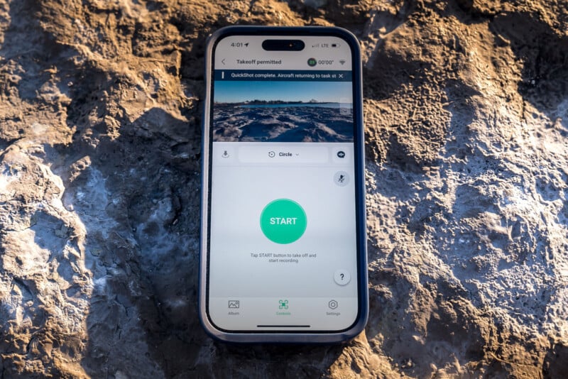 A smartphone displaying a drone control app is resting on a rocky surface. The screen shows a green "START" button and a small aerial image at the top. The device is in a rugged case, capturing natural light.