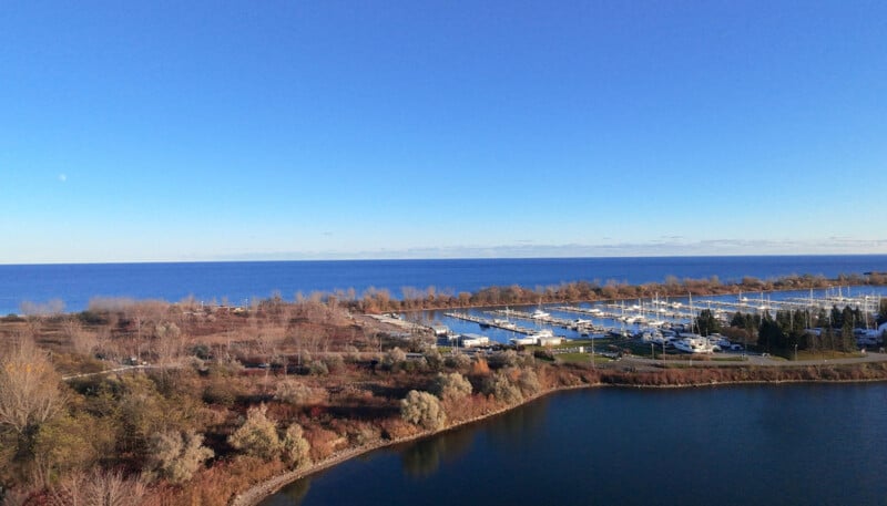 A panoramic view of a marina with several boats docked along the shore. The foreground features calm water and autumn-colored trees. The background showcases a vast expanse of blue sky and the lake meeting the horizon.