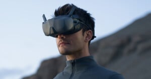 A person wearing dark virtual reality goggles stands outdoors against a blurred mountainous background. They are wearing a dark, high-collared jacket and the image captures a serene, expansive setting in natural light.