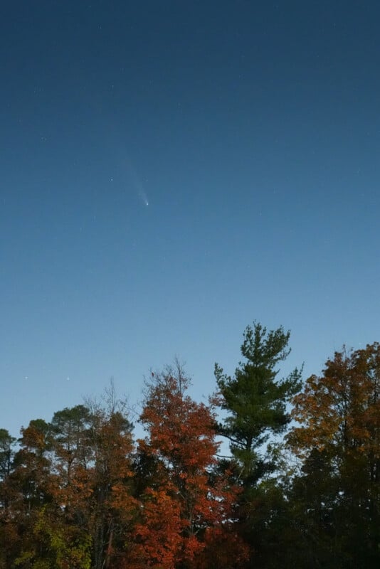 A comet streaks across a clear night sky above a forest with colorful autumn trees.