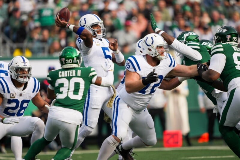 A football game action shot: A quarterback in a white and blue uniform throws a pass while being blocked by players in green uniforms. Teammates and opponents surround him on the field, with the crowd visible in the background.