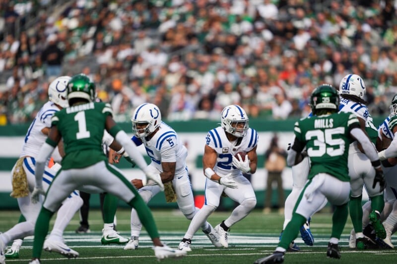 Football game scene with players in blue and white uniforms facing players in green uniforms on a field. A player in blue is running with the ball while teammates and opponents surround him. Stadium crowd is visible in the background.