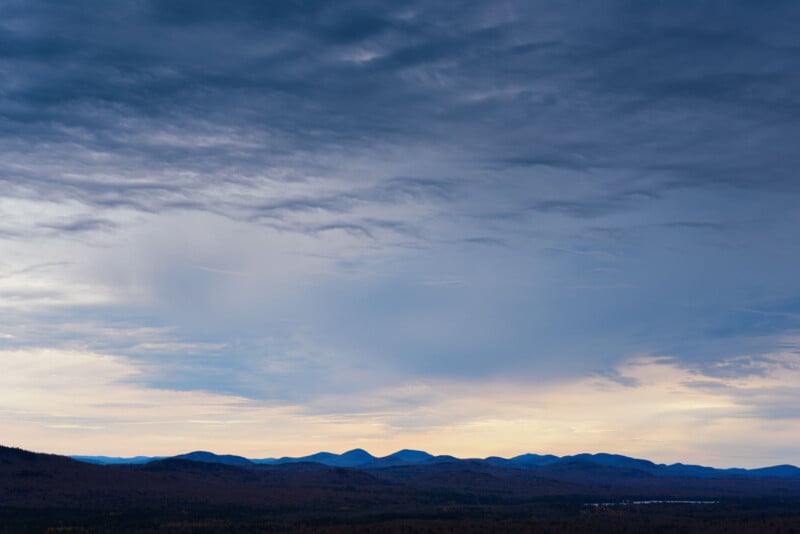 A scenic view of a landscape with rolling hills silhouetted against a dramatic, cloudy sky. The horizon shows a blend of soft, fading daylight transitioning into dusk, creating a tranquil and expansive atmosphere.