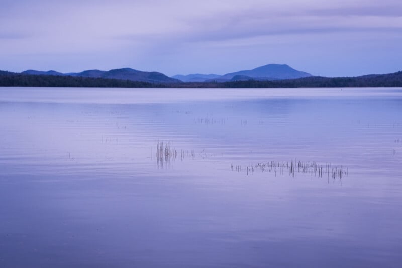 A tranquil lake with a smooth surface reflecting the soft hues of the evening sky. Sparse reeds emerge from the water in the foreground. Rolling hills and mountains line the horizon under an expansive, cloud-streaked sky.