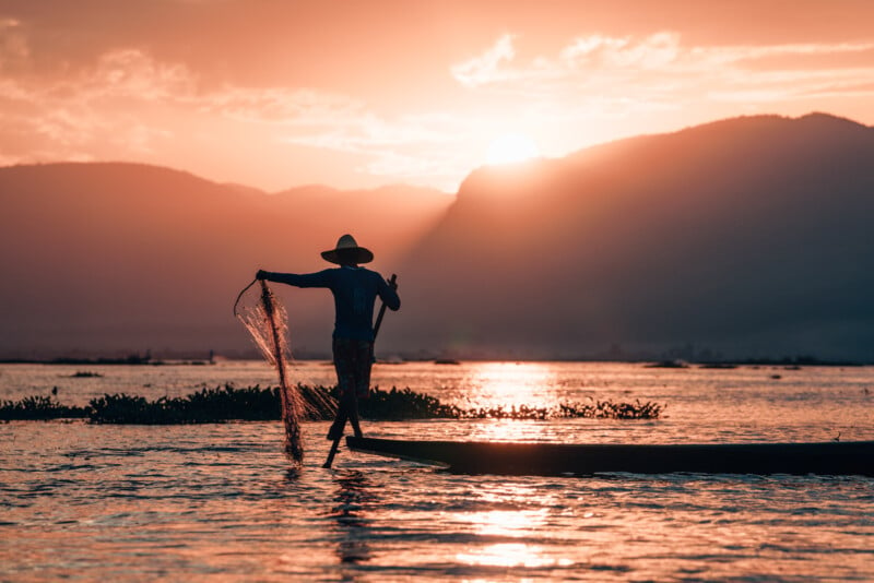 A person in a hat stands on a boat, casting a fishing net into a lake during sunrise or sunset. The sun glows behind distant mountains, reflecting on the water's surface, creating a tranquil and picturesque scene.