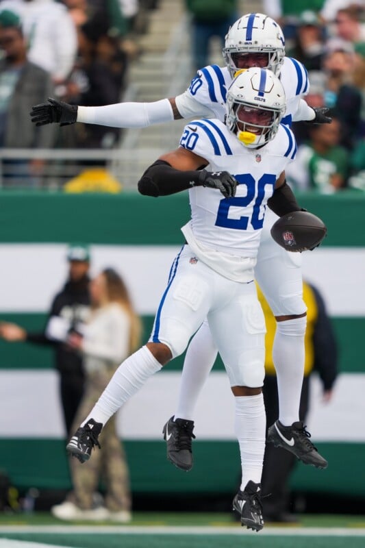 Two football players in blue and white uniforms celebrate on the field. The player in the foreground holds a football, while both appear to be jumping in the air. A blurred crowd is visible in the background.