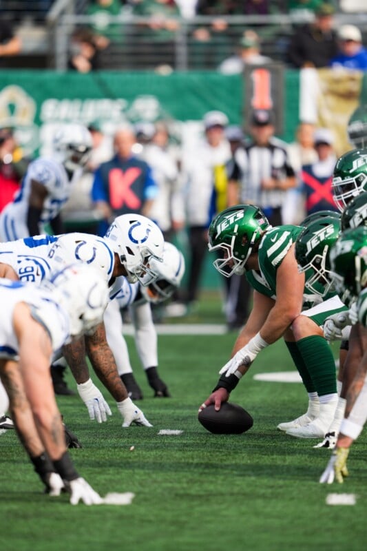 Football players in green and white uniforms face off at the line of scrimmage during a game. A player in green crouches over the ball, preparing to snap it. The field and crowd are visible in the background.