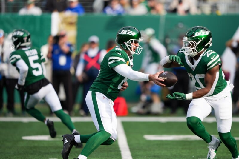 Two football players from the Jets are on the field. One player is handing off the ball to a teammate in a running play. Another player is in the background, running. The game is being played outdoors in a stadium with spectators.