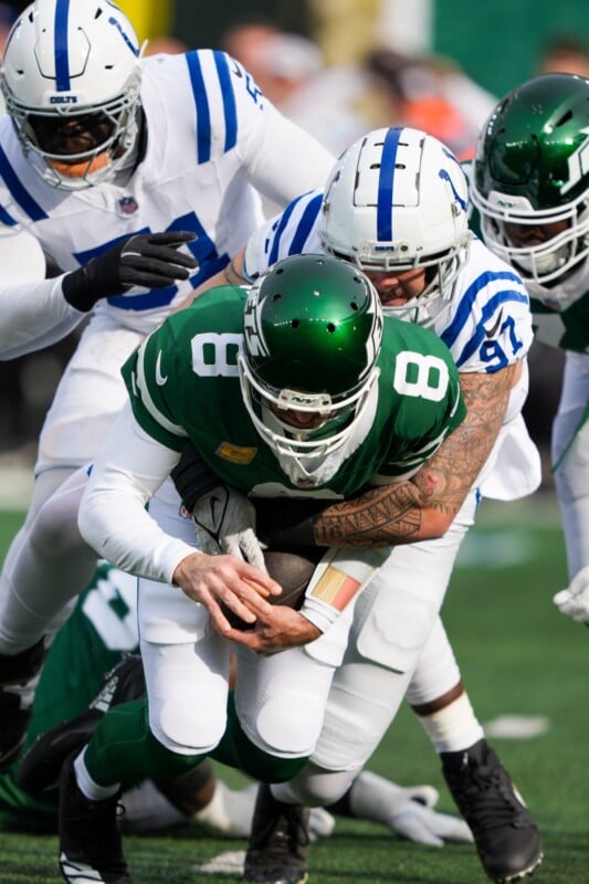 A football player in a green uniform is being tackled by two players in white and blue uniforms during a game. The player in green is holding the ball tightly while falling forward onto the field.