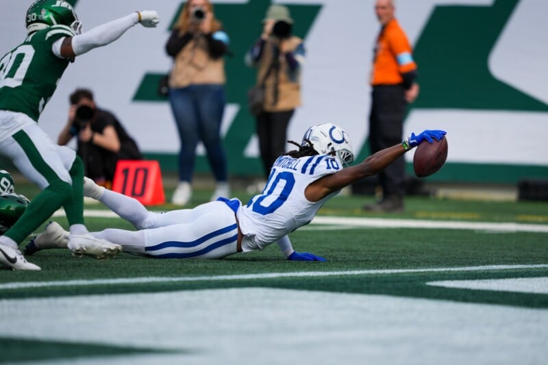 A football player in a white and blue uniform dives with an outstretched arm, holding a football near the end zone. Green-uniformed opposing team players are in pursuit on a grassy field, with a blurred crowd behind.