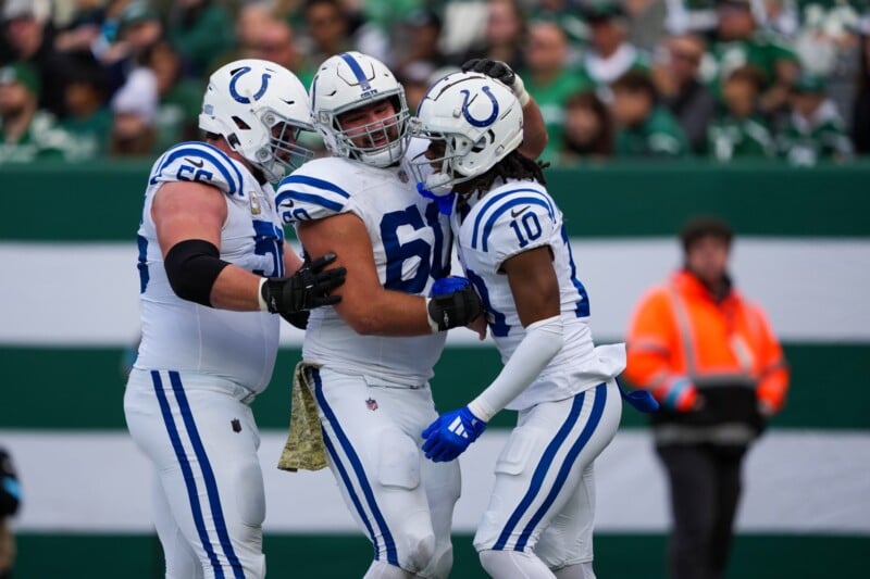 Three football players in white and blue uniforms celebrate on the field, embracing each other. A crowd is visible in the background, with one person in an orange jacket near a striped barrier.