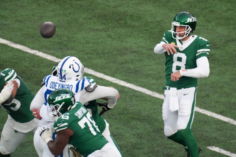 A football player in a green jersey, number 8, throws a pass during a game. Opponents in blue and white uniforms attempt to block. The scene captures the action on a green field.