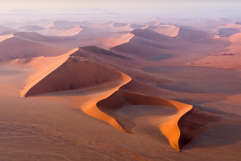 A breathtaking aerial view of vast desert dunes at sunrise, with soft light casting deep shadows and highlighting the undulating sand patterns. The landscape appears endless, with a hazy horizon and distant mountains in the background.