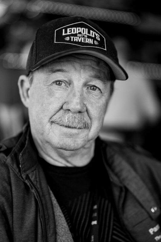 Black and white portrait of an older man with a mustache, wearing a "Leopold's Tavern" hat and a dark jacket. He stares directly at the camera with a neutral expression. The background is softly blurred.