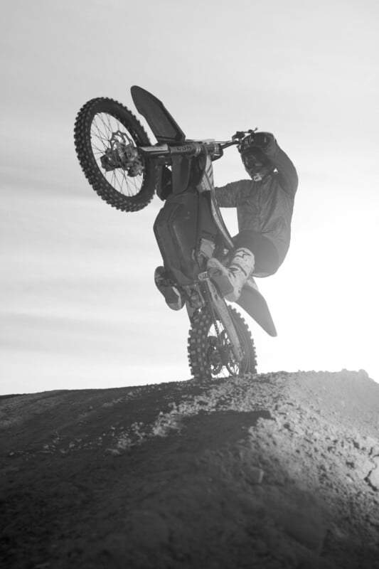 A motorcyclist performs a wheelie on a dirt bike against a cloudy sky backdrop. The grayscale image captures the silhouette of the rider and bike as they ascend a dirt hill.