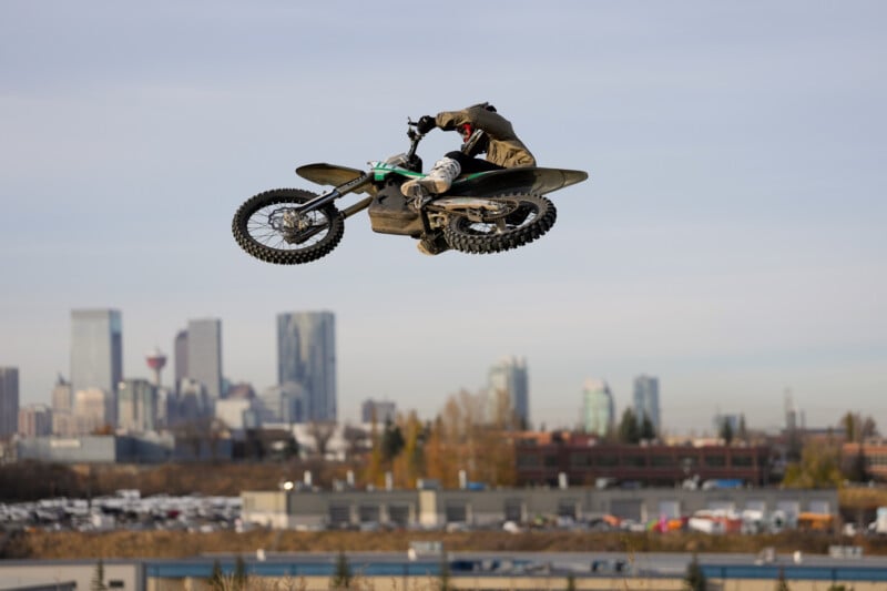 A person performs a high jump on a motocross bike against a city skyline backdrop with tall buildings and autumn foliage in the foreground.