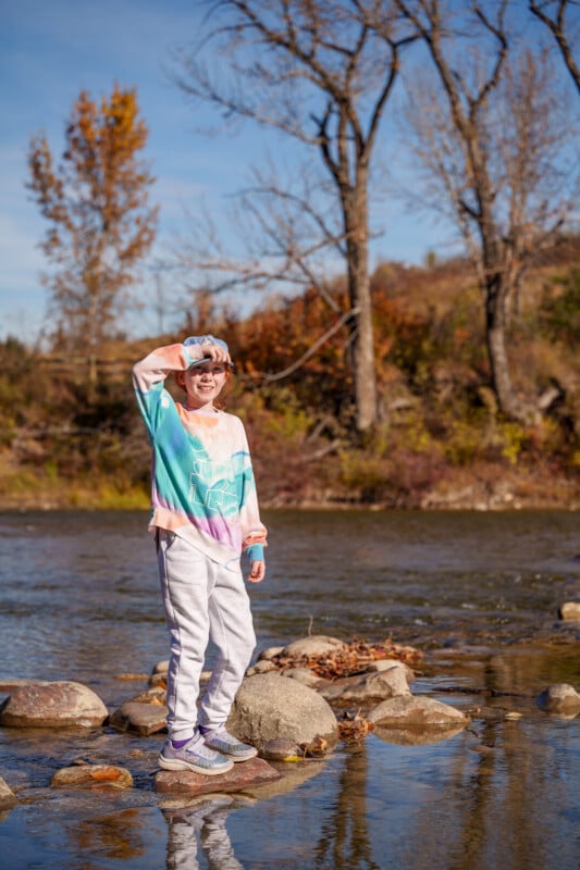 A child in a colorful sweatshirt and gray pants stands on rocks by a river, holding a camera to their eye. The background features bare trees and a clear blue sky on a sunny day.