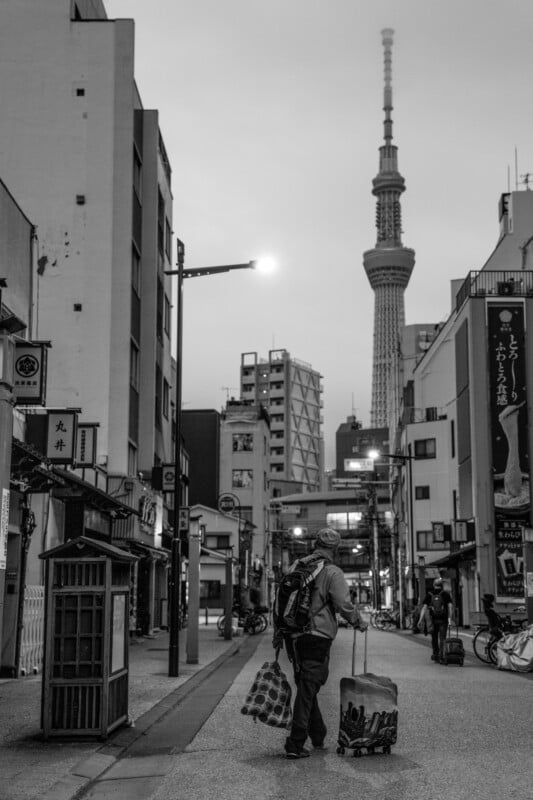 A black and white photo of a person walking down a street in Tokyo, carrying shopping bags. The Tokyo Skytree towers in the background. The street is lined with buildings, bicycles, and a phone booth.