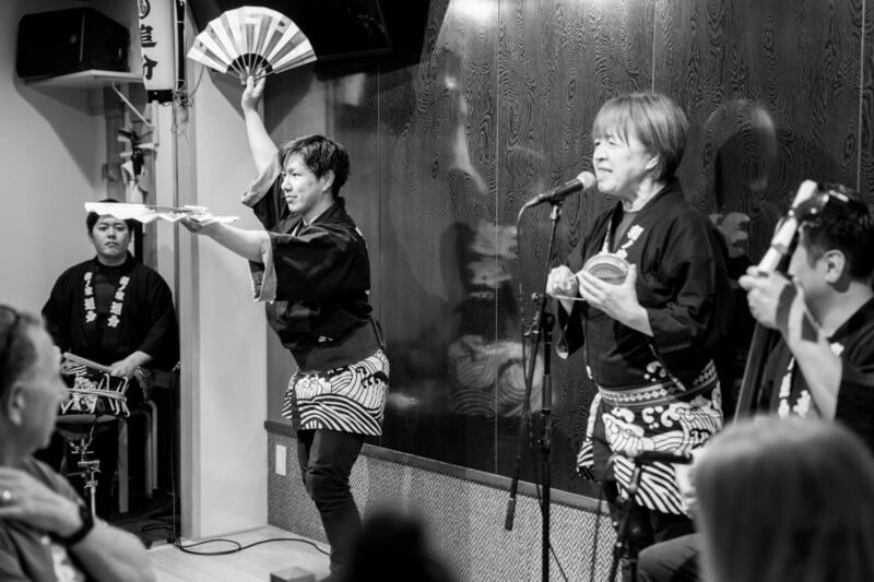 A black and white photo of a live performance. Performers wear traditional clothing; one plays a drum, another holds a fan, and a third sings into a microphone. An audience watches attentively.