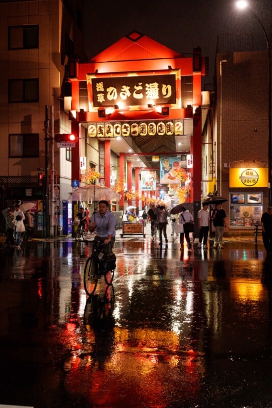 A bustling city street at night with a cyclist riding through a wet, reflective surface. Neon lights illuminate the scene, and several people walk under the brightly lit entrance of a shopping area with Japanese signage.