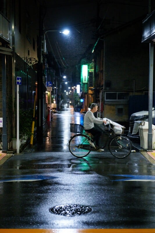 A person is riding a bicycle on a wet street at night, with reflections from streetlights shining on the road. The scene is dimly lit, and the background features a narrow, rain-soaked alley with various buildings and signs.