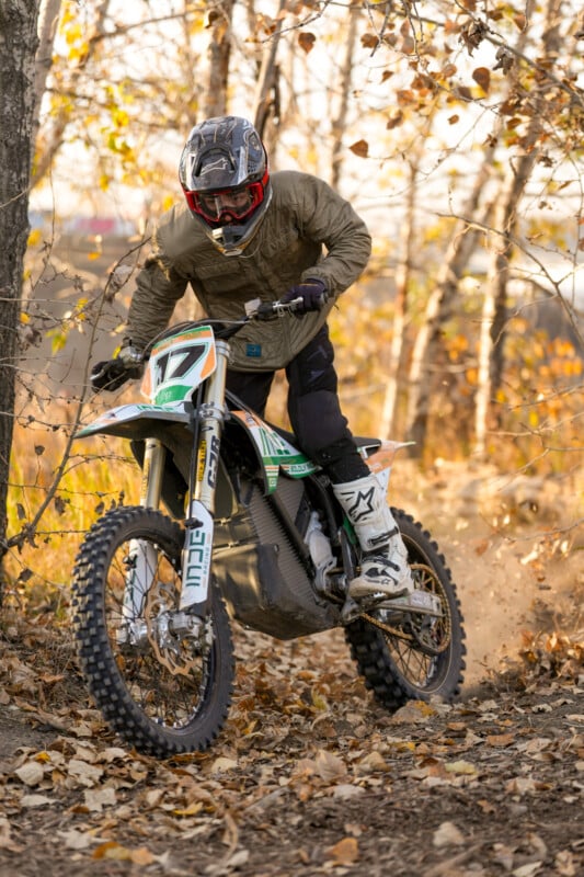 A person rides a dirt bike with the number 17 through a wooded area covered in fallen leaves. They are wearing a helmet and protective gear. The background features leafless trees and soft evening sunlight filtering through.