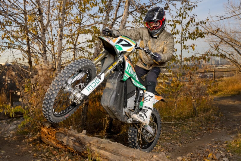 A person in protective gear rides a dirt bike over a log in a wooded area. The bike is airborne, and fallen leaves are scattered on the ground. The rider appears to be at a dirt biking trail with autumn foliage in the background.