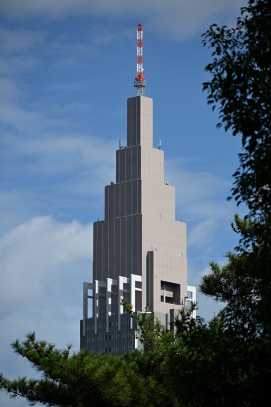 The image shows a tall, modern skyscraper with a stepped design against a partly cloudy blue sky. Tree branches are visible in the foreground.