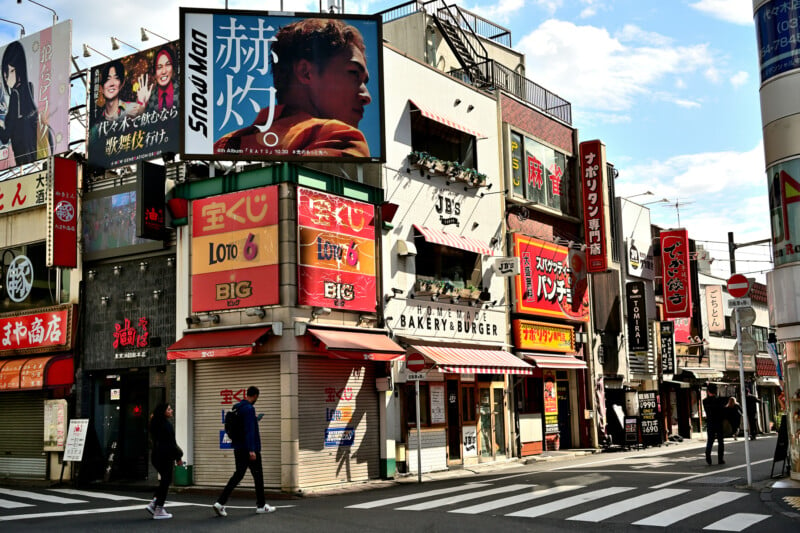 A busy urban street scene in Japan with colorful storefronts and billboards. Pedestrians walk along the sidewalk under a bright sky. Prominent signs advertise various shops, including a burger place and a convenience store.