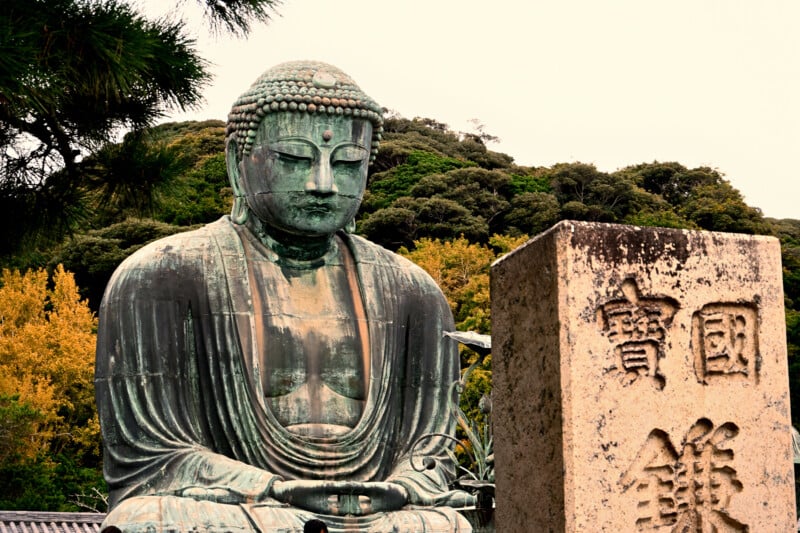 The image shows a large bronze statue of Buddha, seated outdoors against a backdrop of trees. There is a stone plaque with inscriptions in front of the statue. The scene conveys a calm and serene atmosphere.