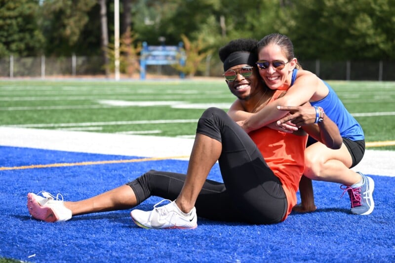 Two athletes sitting on a blue track, smiling and hugging each other. One wears an orange shirt and sunglasses, the other in a blue tank top and sunglasses. Green field and trees in the background.