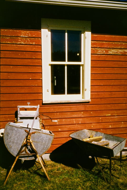 A red wooden wall with a white-framed window. In front, there's an old metal washing machine and a wheelbarrow. Grass is visible on the ground. The scene is lit by warm sunlight.